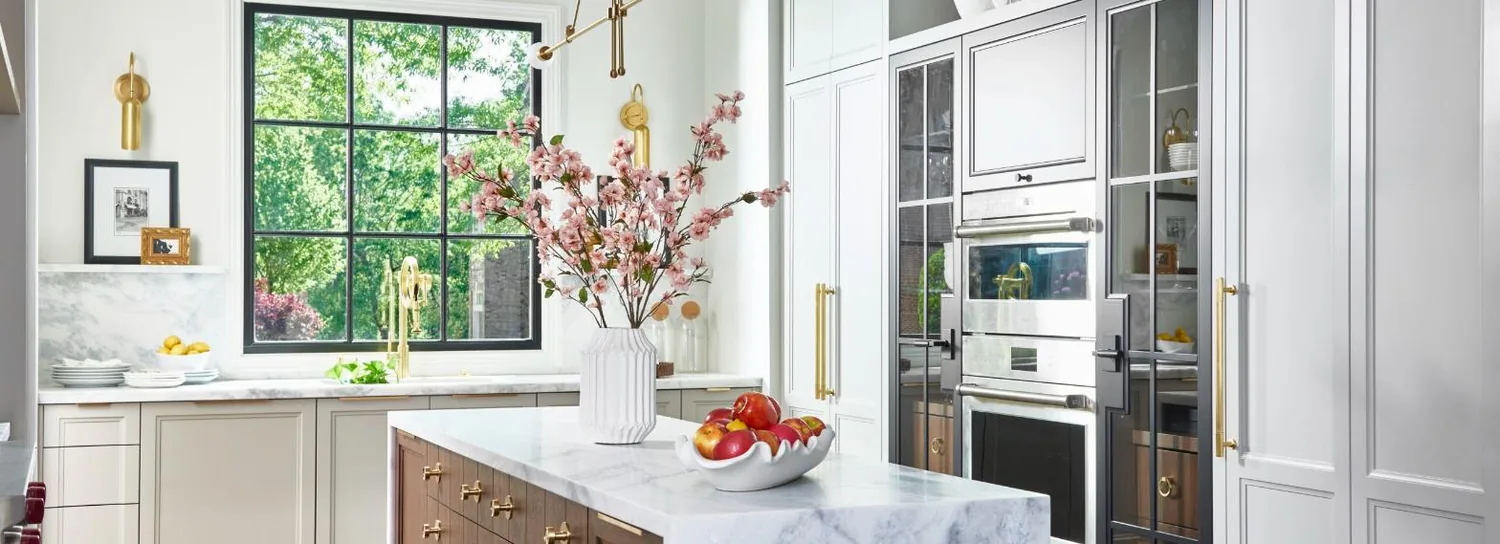 Wide view of the Lakehaus kitchen: brass faucet at a steel-framed window, marble island and warm wood cabinetry